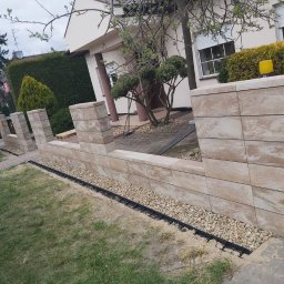 MAX-BUD - Nowo wybudowany murek z beżowych kostek brukowych, oddzielający trawnik od chodnika, z widoczną czarną rynną odprowadzającą wodę i żółtym kontenerem na gruz w tle.