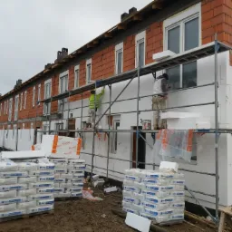 Dwóch pracowników na rusztowaniu przy ocieplaniu elewacji szeregu domów z czerwonej cegły, widoczne paczki styropianu i worki z zaprawą.