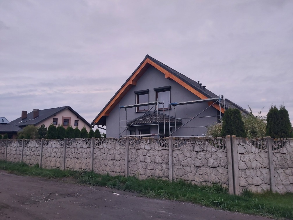 Dom jednorodzinny w trakcie malowania elewacji na szaro, widoczne rusztowania i fragment ozdobnego betonowego ogrodzenia.