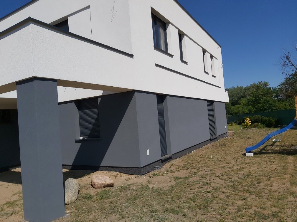 Nowoczesny dom jednorodzinny z białą elewacją i szarą podmurówką w Rogoźnie, widok z boku, na trawniku leżą kamienie, w tle zieleń drzew.