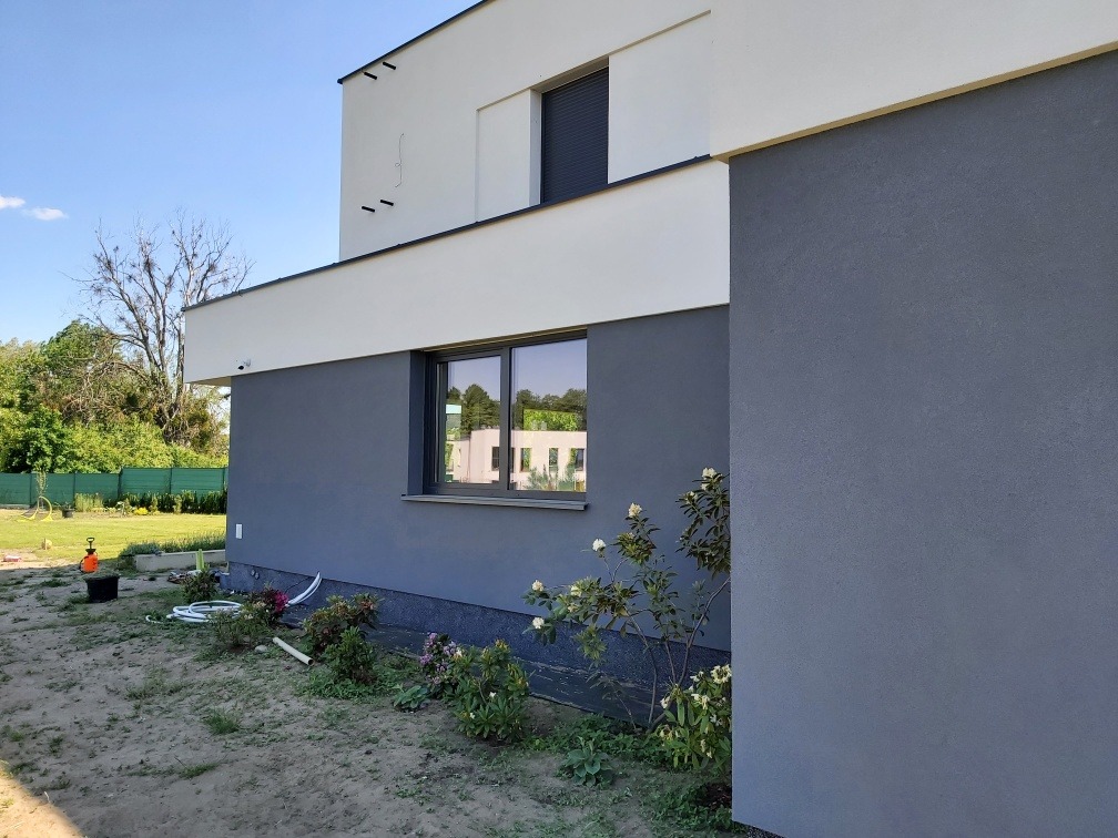 Nowoczesny dom z szarą elewacją, widok z boku, fragment ogrodu z młodymi roślinami i sprzętem ogrodniczym.