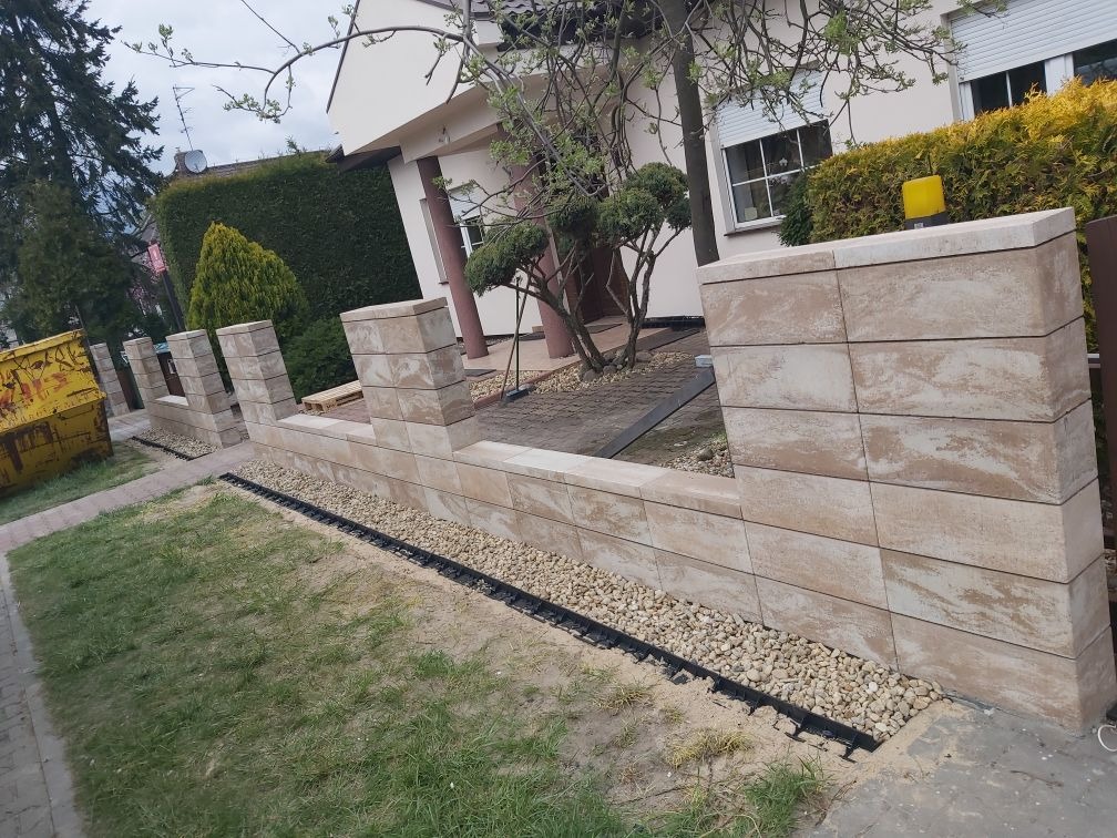 Nowo wybudowany murek z beżowych kostek brukowych, oddzielający trawnik od chodnika, z widoczną czarną rynną odprowadzającą wodę i żółtym kontenerem na gruz w tle.