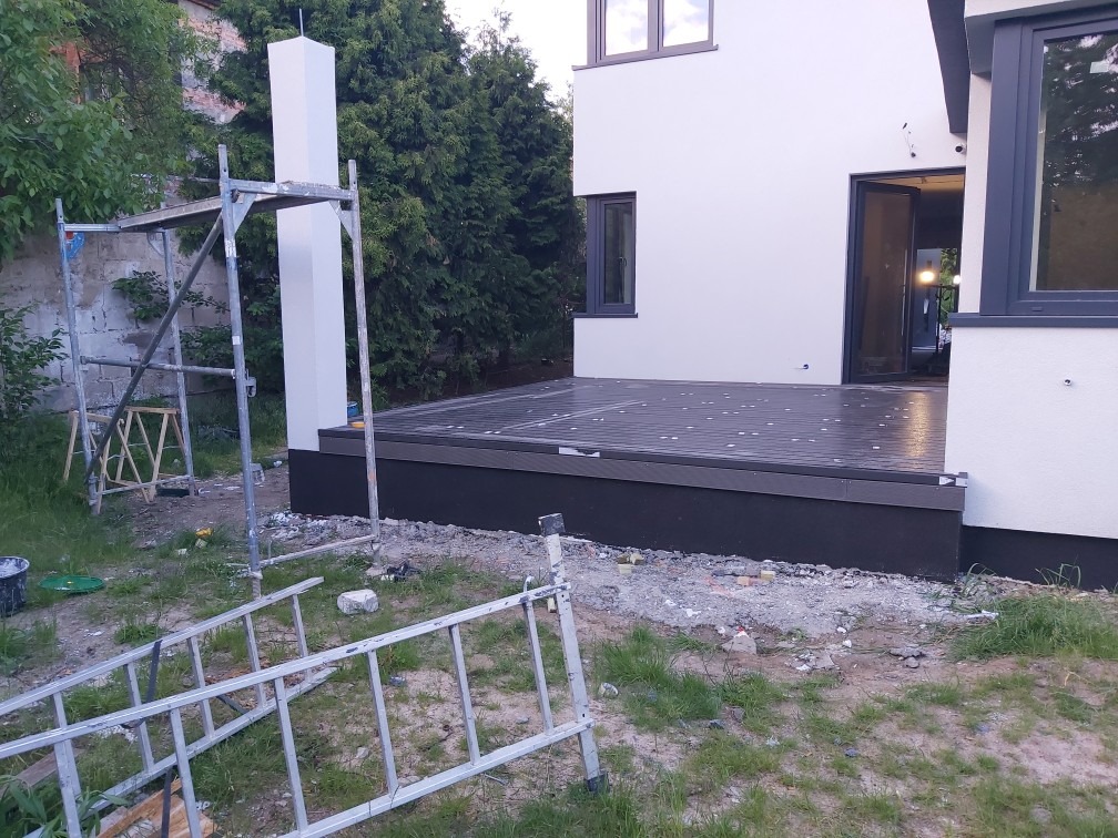 Nowoczesny dom z jasną elewacją i tarasem z kompozytowych desek, widoczne rusztowanie oraz teren w trakcie prac wykończeniowych.