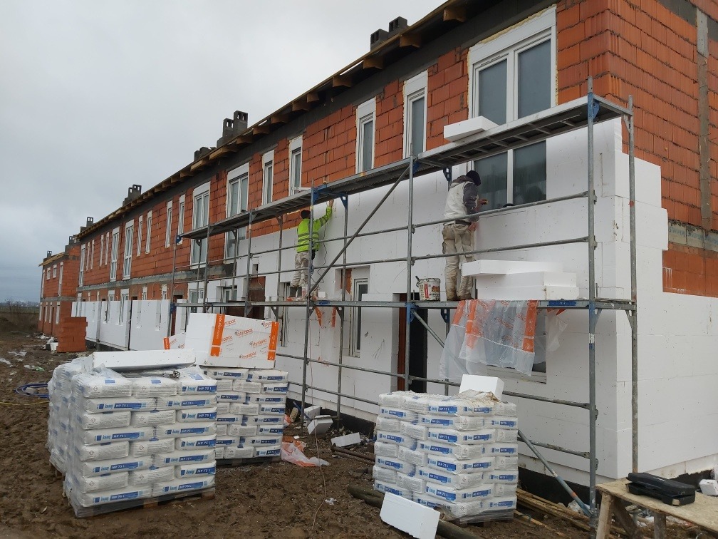 Dwóch pracowników na rusztowaniu przy ocieplaniu elewacji szeregu domów z czerwonej cegły, widoczne paczki styropianu i worki z zaprawą.