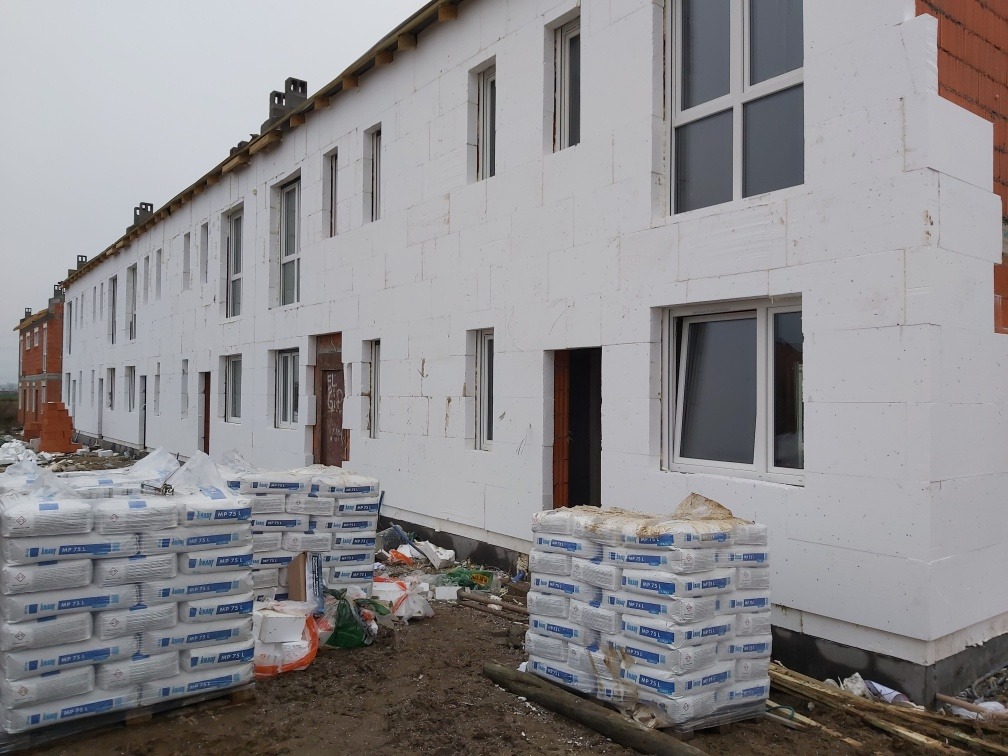 Budynek w trakcie ocieplania styropianem, widoczne worki z zaprawą klejową MP 75L, surowy stan elewacji i okien.