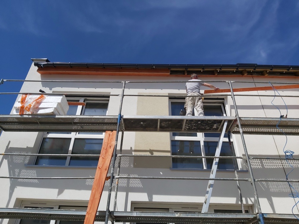 Pracownik na rusztowaniu montuje elementy konstrukcyjne na elewacji budynku, widoczne płyty styropianowe i przygotowane fragmenty do mocowania.