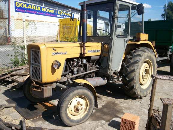 Żółty ciągnik rolniczy Ursus C-360 3P z przyczepą w tle, na tle budynku z szyldem Solaria Nowe i Używane.
