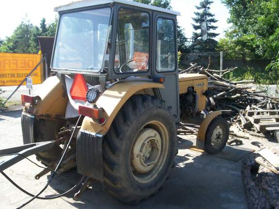 Żółty ciągnik rolniczy Ursus C 360 3P z widocznymi śladami użytkowania, zaparkowany na betonowym placu obok sterty gałęzi i palet.