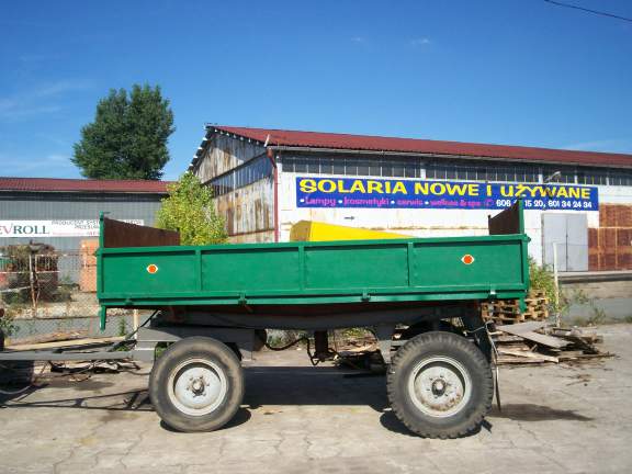 Zielona przyczepa rolnicza Sanok D 47A z widocznymi oponami i elementami konstrukcyjnymi, zaparkowana na betonowym placu przed budynkiem z reklamą solarium.