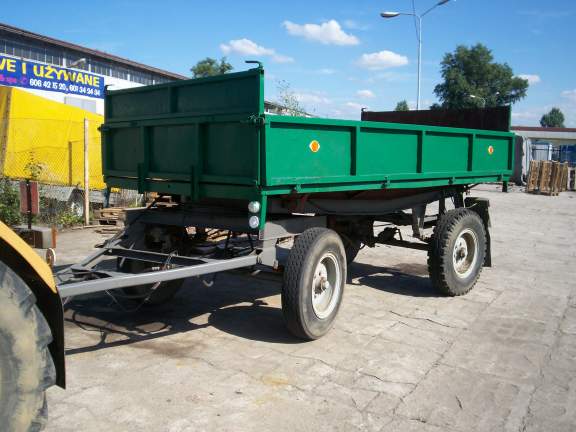 Zielona przyczepa rolnicza Sanok D 47A z widocznymi oponami i konstrukcją podwozia na betonowym placu, w tle budynek z szyldem i żółta plandeka.