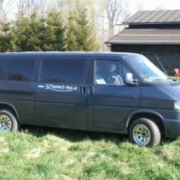 Czarny bus marki Volkswagen Transporter T4 z napisem 'KARPACZ-BUS, przewóz osób' na boku, zaparkowany na trawie przed budynkiem gospodarczym w słoneczny dzień.