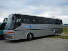 Srebrny autokar z napisem 'KARPACZ-BUS, przewóz osób' na boku, zaparkowany na żwirowym parkingu przy pochmurnym niebie.