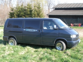 Czarny bus marki Volkswagen Transporter T4 z napisem 'KARPACZ-BUS, przewóz osób' na boku, zaparkowany na trawie przed budynkiem gospodarczym w słoneczny dzień.