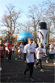 Uczestnicy biegu ulicznego z numerami startowymi, na tle dmuchanych reklam i drzew bez liści, w słoneczny dzień.