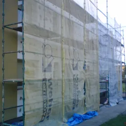 Budynek w trakcie ocieplania elewacji, osłonięty siatką z nadrukiem reklamowym, widoczne rusztowania i fragment chodnika z niebieską folią zabezpieczającą.