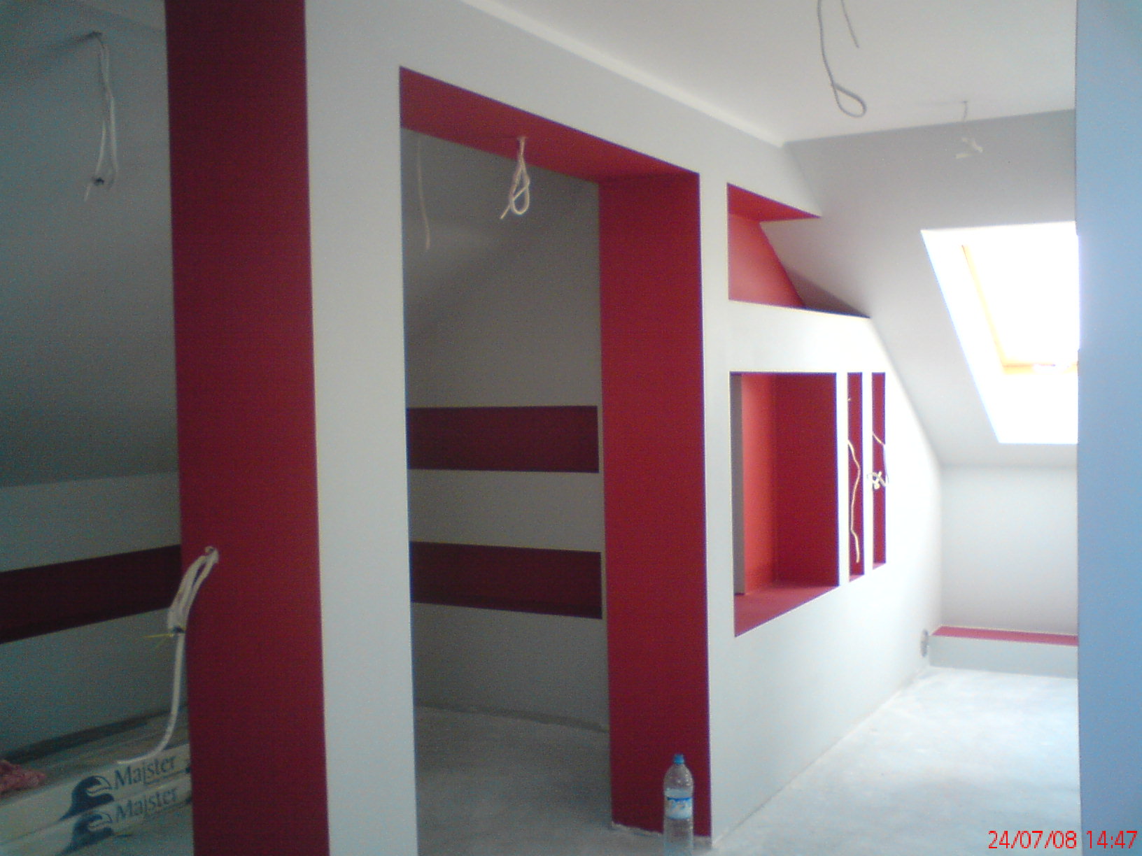 Wnętrze poddasza w trakcie wykańczania; widoczne ściany pomalowane na biało z elementami dekoracyjnymi w kolorze czerwonym, wnęki w ścianach, okno dachowe, przewody elektryczne zwisające z sufitu...