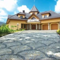 Elegancki podjazd do domu wyłożony ozdobną nawierzchnią z drobnej kostki w odcieniach szarości, ułożonej we wzór wachlarzy. W tle widoczny duży, dwupiętrowy dom z garażem i wieżyczką.