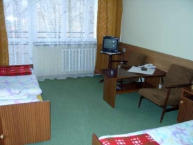 Pokój w sanatorium w Iwoniczu Zdroju z dwoma pojedynczymi łóżkami z czerwonymi narzutami, stolikiem kawowym, telewizorem i fotelem. Widok na okno z zasłonami.