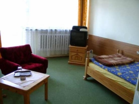 Pokój w ośrodku wczasowym: pojedyncze łóżko z niebieską pościelą w kwiaty, mały stolik nocny z telewizorem, czerwony fotel i stolik kawowy z pilotem, zielona wykładzina.