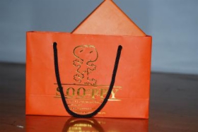 Pomarańczowa torba papierowa z czarnymi uchwytami, zdobiona złotym nadrukiem postaci z kreskówki i napisem 'SOO PET'. W torbie widoczny fragment pomarańczowego pudełka.