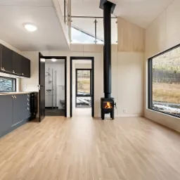 Wnętrze domu z jasną drewnianą podłogą, czarną kuchnią z drewnianym blatem, kominkiem i dużym oknem z widokiem na góry.