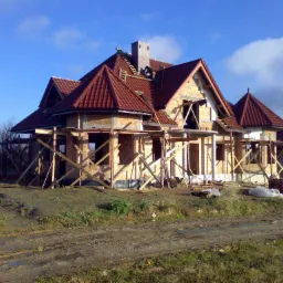 Wznoszony dom z czerwonej cegły i dachówką w trakcie prac dekarskich, widoczny murarz na dachu, drewniane rusztowania wokół budynku i błotnista droga dojazdowa.