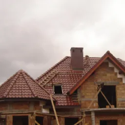 Niedokończony dom murowany z cegły z czerwoną dachówką, widoczne rusztowania i drabina na dachu, pochmurne niebo w tle.