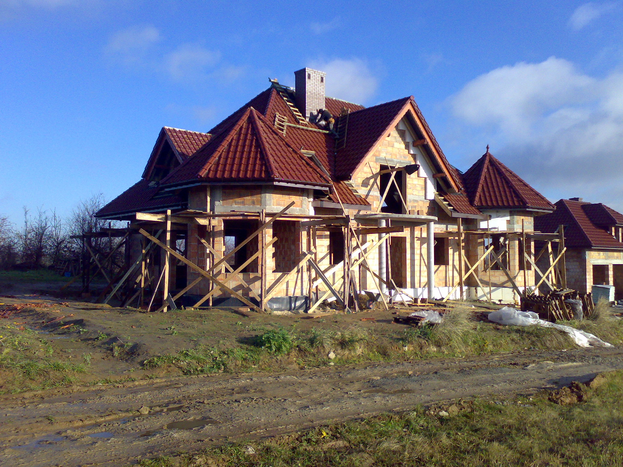 Wznoszony dom z czerwonej cegły i dachówką w trakcie prac dekarskich, widoczny murarz na dachu, drewniane rusztowania wokół budynku i błotnista droga dojazdowa.