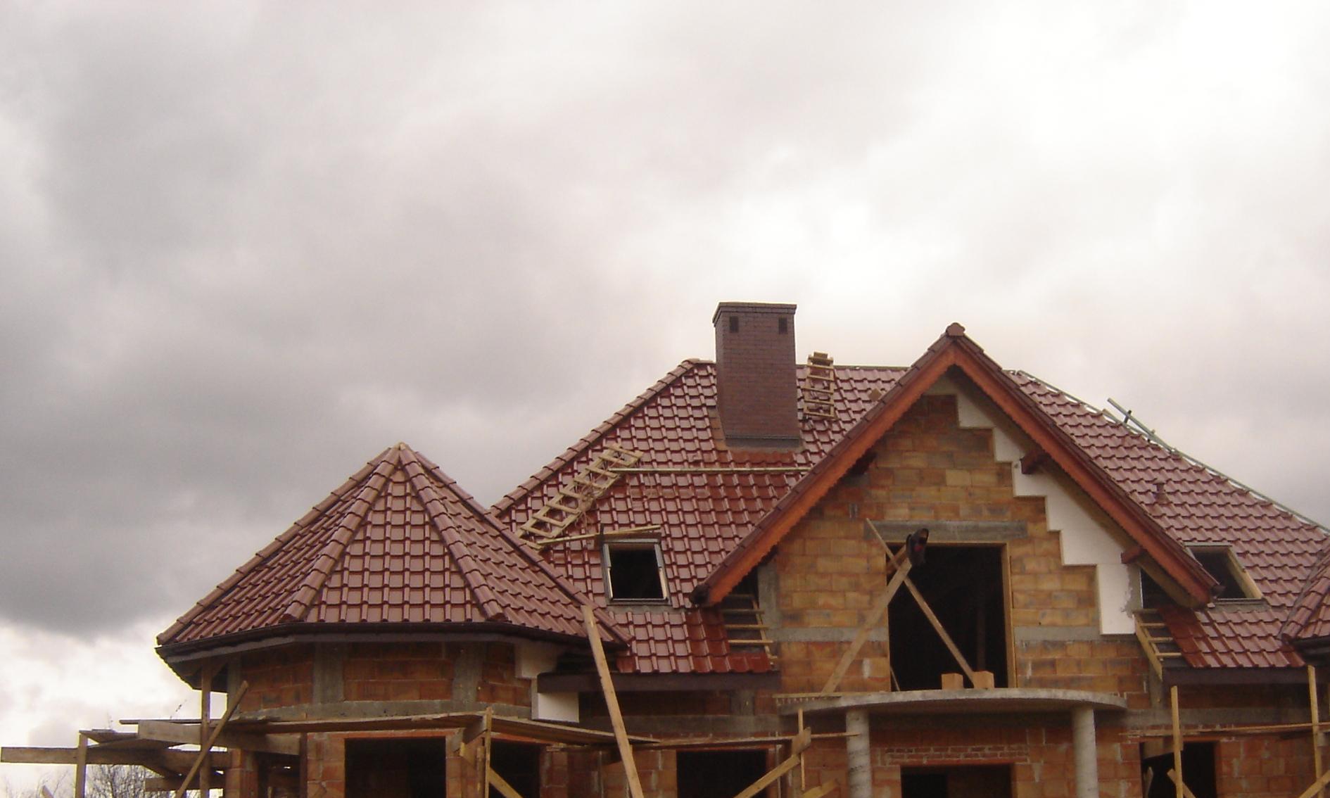 Niedokończony dom murowany z cegły z czerwoną dachówką, widoczne rusztowania i drabina na dachu, pochmurne niebo w tle.