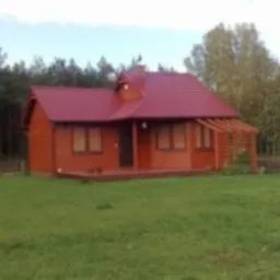 Mały, brązowy domek z czerwonym dachem na zielonej łące, otoczony drzewami. Widoczna antena satelitarna na dachu. Dom z drewna.