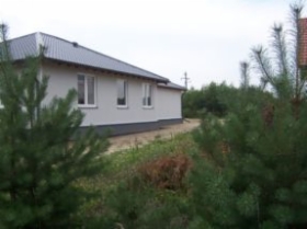 Dom jednorodzinny w trakcie rozbudowy w miejscowości Cała Polska, widok zza drzew iglastych, ściany otynkowane na biało, szary dach.