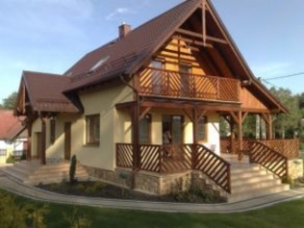 Jasny, parterowy dom z poddaszem użytkowym, drewnianą konstrukcją balkonu i zadaszenia nad wejściem, brązowy dach, kamienne wykończenie elewacji na dole, drewniane schody wejściowe.