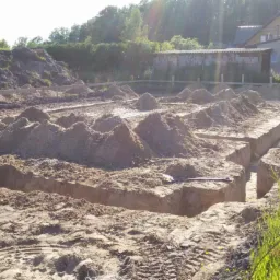 Wykopy pod fundamenty z widocznym zbrojeniem w rowach, przygotowane do zalania betonem. Widoczne drewniane szalunki i pryzmy ziemi. Słoneczny dzień.
