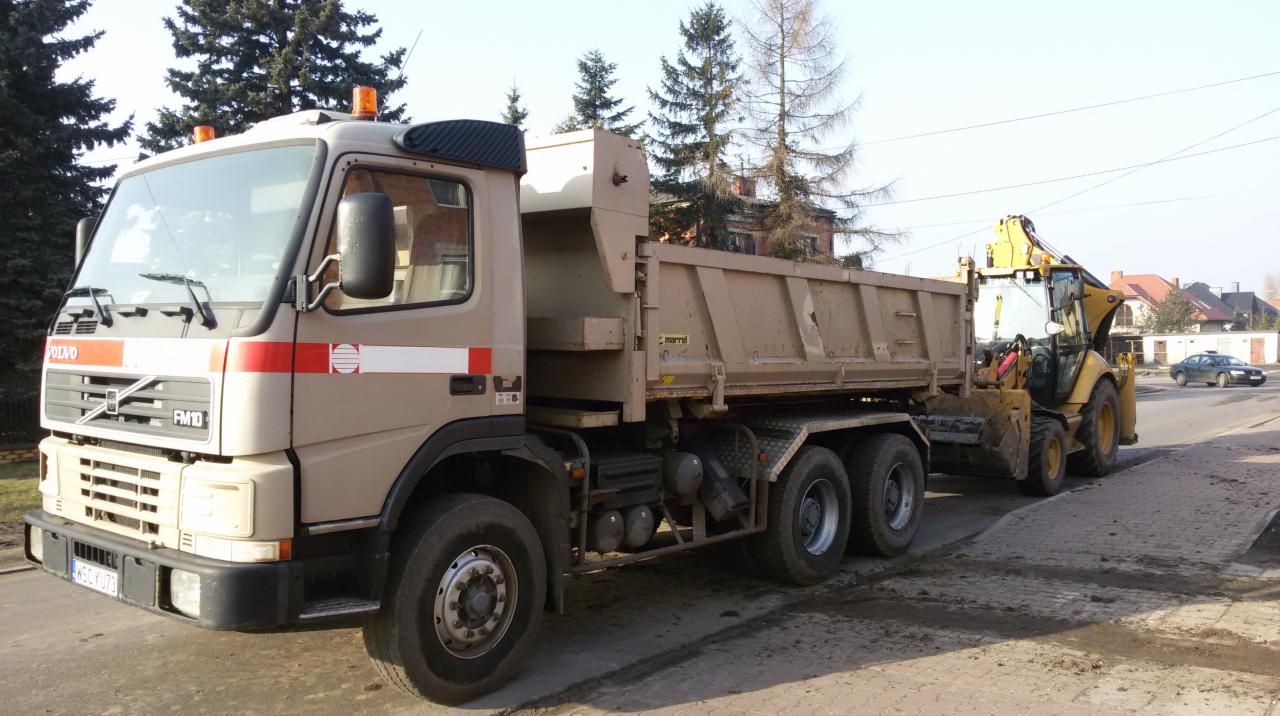 Ciężarówka Volvo FM10 z żółtą koparko-ładowarką w tle, na tle drzew i budynków. Sprzęt przy pracy na drodze. Widoczne ślady ziemi.