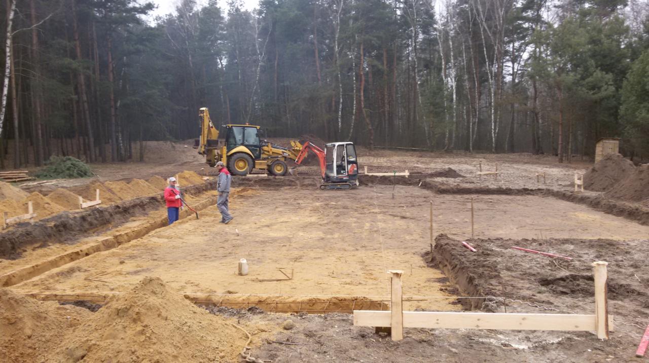 Wykop pod fundamenty domu w lesie, widoczne szalunki i zbrojenia, pracownicy oraz maszyny budowlane: koparka i traktor z łyżką. Etap przygotowania do wylewania fundamentów.