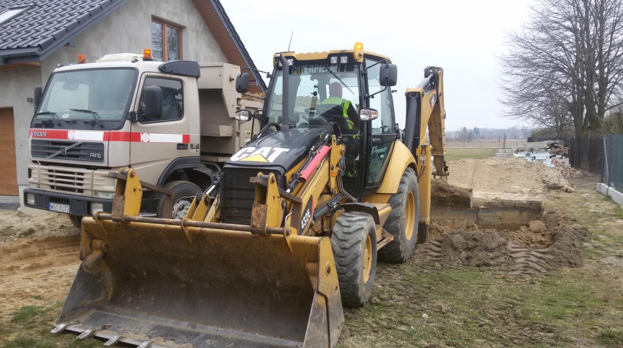 Żółta koparko-ładowarka CAT 432E i ciężarówka Volvo FM10 na działce z wykopem. Operator w kabinie. Częściowo widoczny budynek w tle.