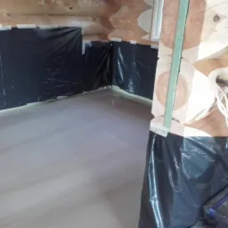 Świeżo wylana, gładka posadzka jastrychowa w drewnianym domu z bali, zabezpieczona folią przy ścianach, widoczny fragment drewnianej kolumny z przewodami.