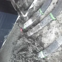 Świeżo wylana, szara masa podłogowa pokrywająca rury ogrzewania podłogowego z zielonymi opaskami, widoczna folia izolacyjna przy ścianie.