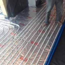 Rozłożone rury ogrzewania podłogowego na folii z siatką, z widocznymi fragmentami osoby w jeansach i brązowych butach, oraz zabezpieczonym słupem konstrukcyjnym