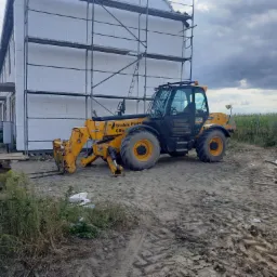 Żółta ładowarka teleskopowa JCB na tle budynku w trakcie ocieplania styropianem, widoczne rusztowanie i palety z materiałem budowlanym.