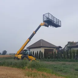 Żółty podnośnik teleskopowy JCB z platformą roboczą na tle budynków jednorodzinnych, widoczne panele słoneczne na dachu jednego z budynków.