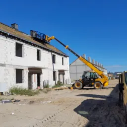 Budowa domu z białego bloczka: żółty podnośnik koszowy przy dachu z położoną dachówką, widoczna żółta furgonetka i inne budynki w tle, słoneczny dzień.