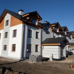 Nowoczesne budynki wielorodzinne z drewnianymi elementami wykończeniowymi pod dachem, czarną dachówką i szarą elewacją w trakcie budowy, widoczne materiały budowlane i teren budowy.