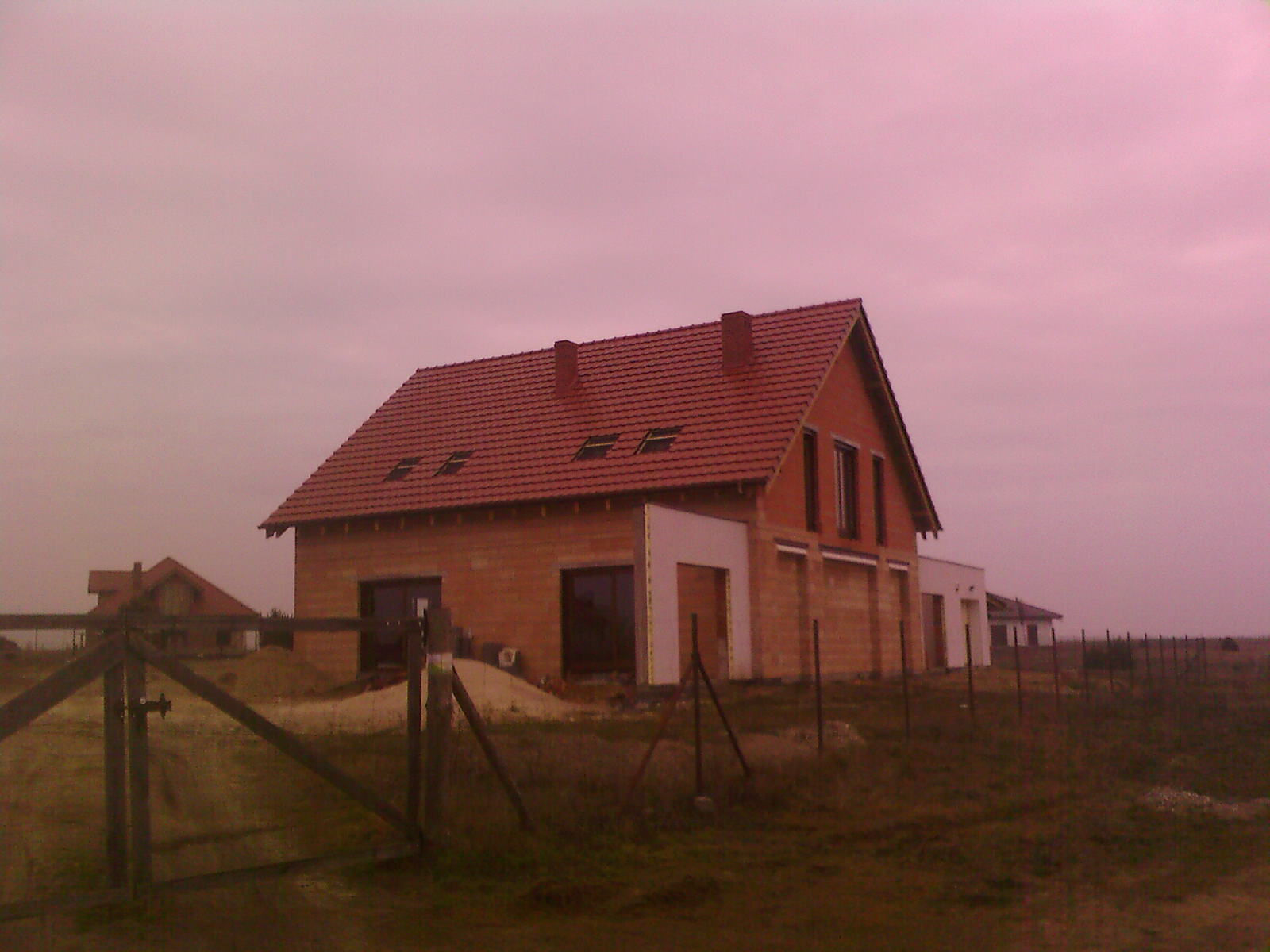 Niedokończony dom z czerwonej cegły z ciemnoczerwoną dachówką, widoczne okna dachowe i kominy, ogrodzenie z drewnianych słupków na pierwszym planie, krajobraz polny w tle.