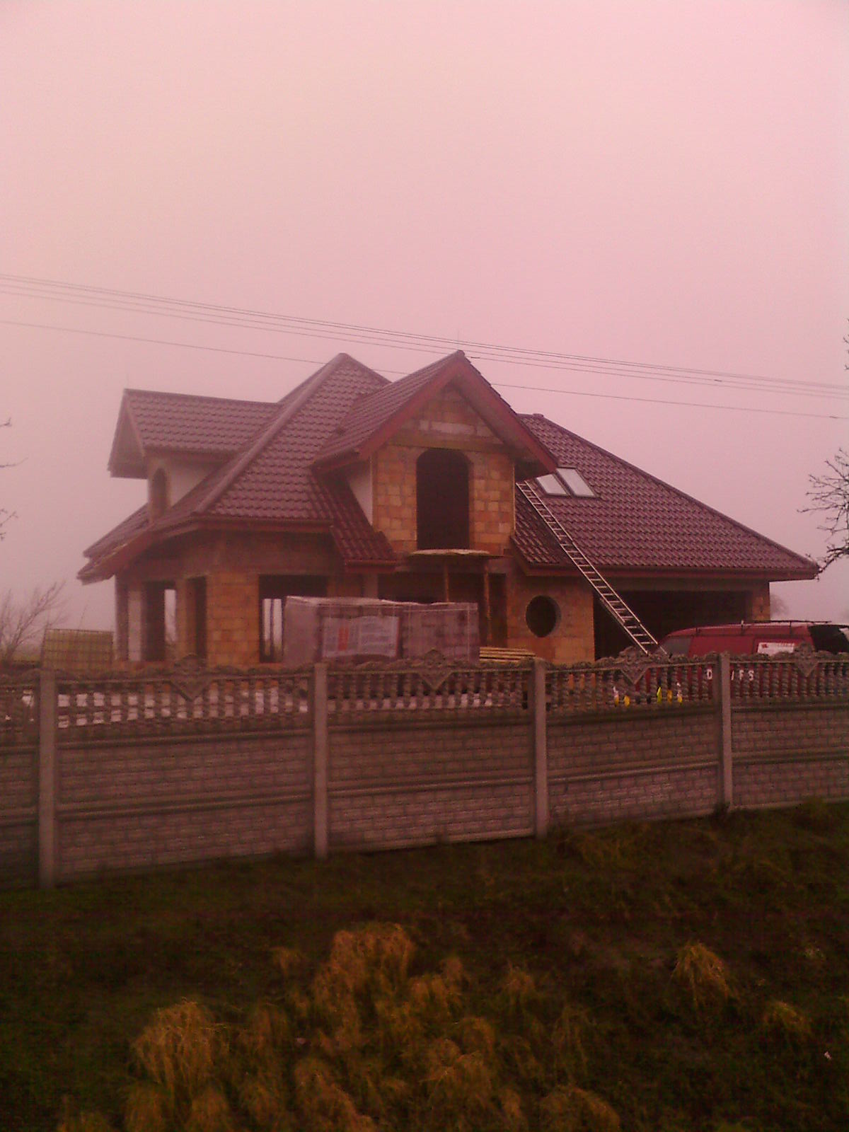 Niedokończony dom z cegły z ciemnoczerwoną dachówką, drabina oparta o dach, widok zza betonowego ogrodzenia w mglisty dzień.