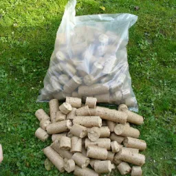 Owalne pale z trocin drzewnych, część wysypana z przezroczystego worka na zielonej trawie, dłoń trzyma jeden z paliw.