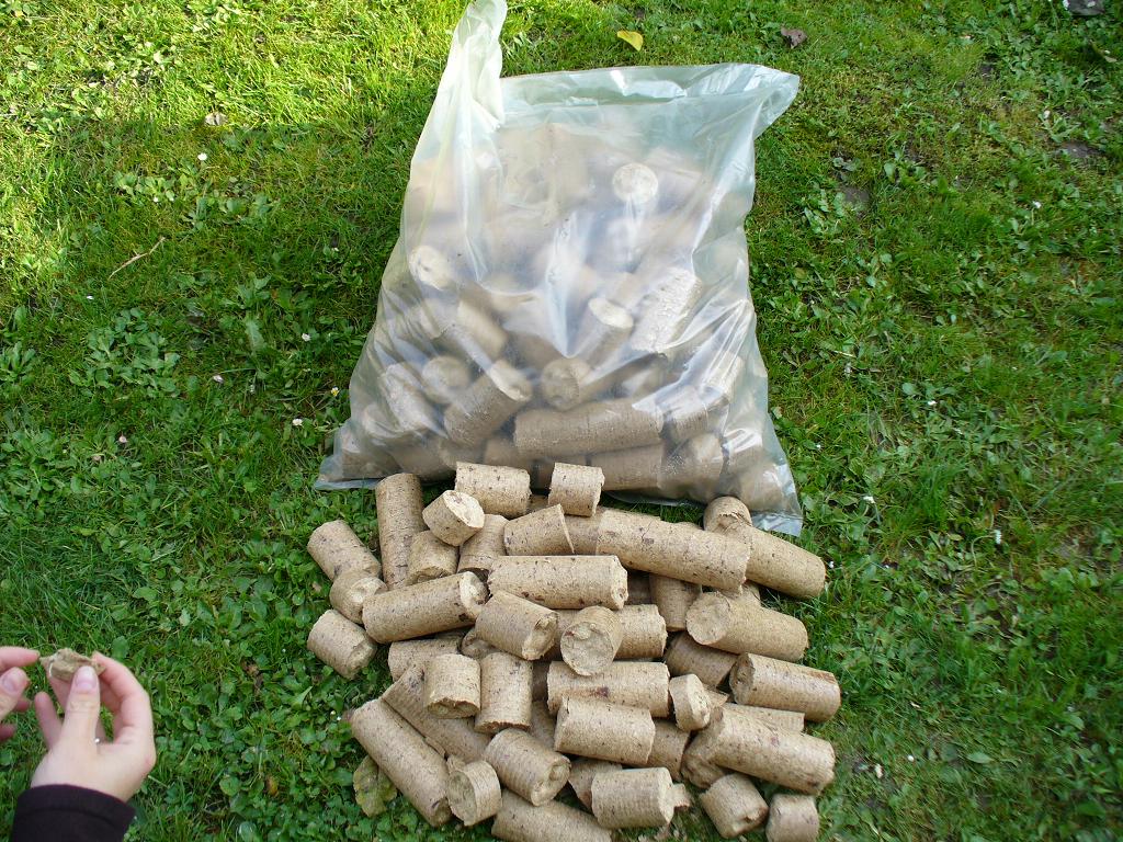 Owalne pale z trocin drzewnych, część wysypana z przezroczystego worka na zielonej trawie, dłoń trzyma jeden z paliw.