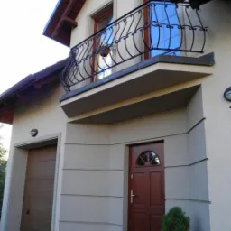 Elewacja domu z balkonem z ozdobną, kutą balustradą, brązowe drzwi wejściowe z półokrągłym przeszkleniem i brama garażowa w tym samym kolorze.