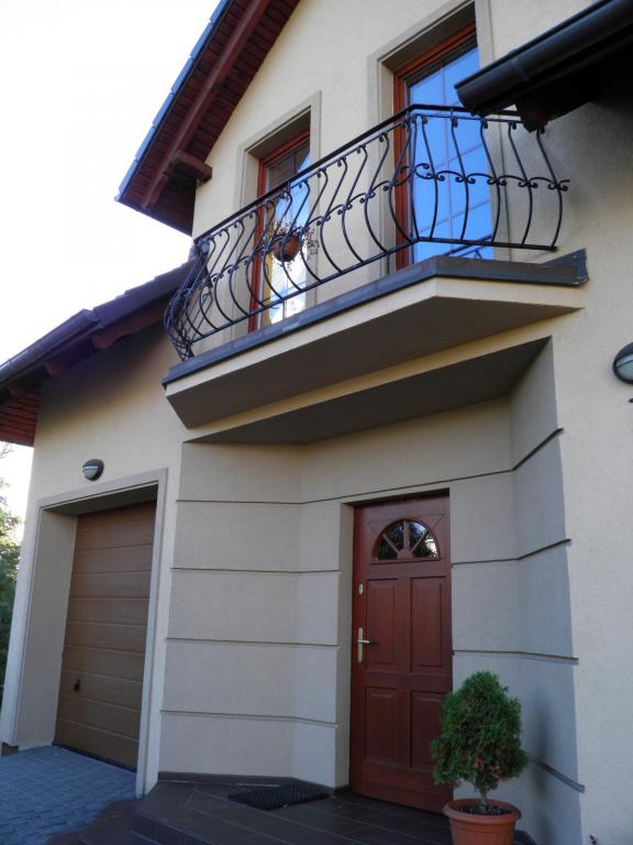 Elewacja domu z balkonem z ozdobną, kutą balustradą, brązowe drzwi wejściowe z półokrągłym przeszkleniem i brama garażowa w tym samym kolorze.