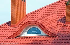 Czerwony dach z blachy na budynku z oknem mansardowym i ceglanym kominem, widoczne regularne wzory tłoczenia na blasze.